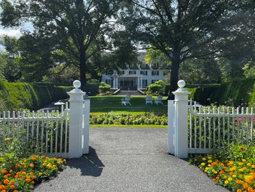 A picturesque view of a mansion garden with a white fence, vibrant flowers, and lush greenery.