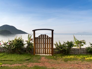 Stone fence with green bushes and grass along and wooden gate to seashore with picturesque view on calm sea with mountain on horizon and blue sky in sunny summer day