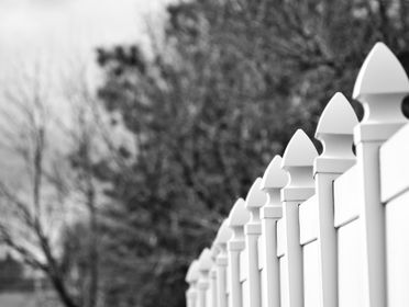 Black and white image of a row of decorative fence posts with blurred winter trees in the background.