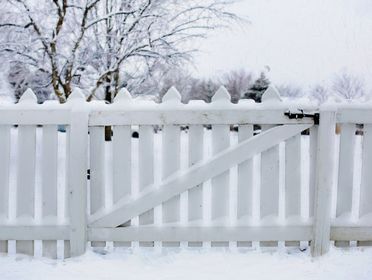 A snow-covered white picket fence in a serene winter landscape, surrounded by frosted trees.