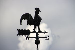 A rooster weathervane silhouetted against a cloudy sky, depicting directions with letters E and W.